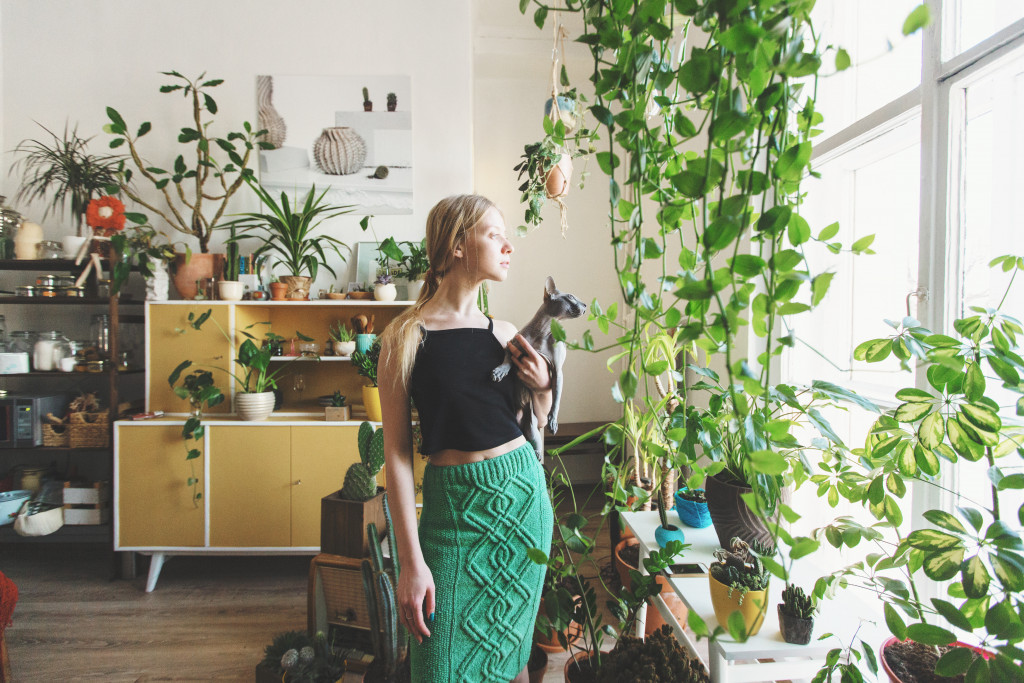 Young woman looking outside a window while holding a cat with indoor plants in the background.